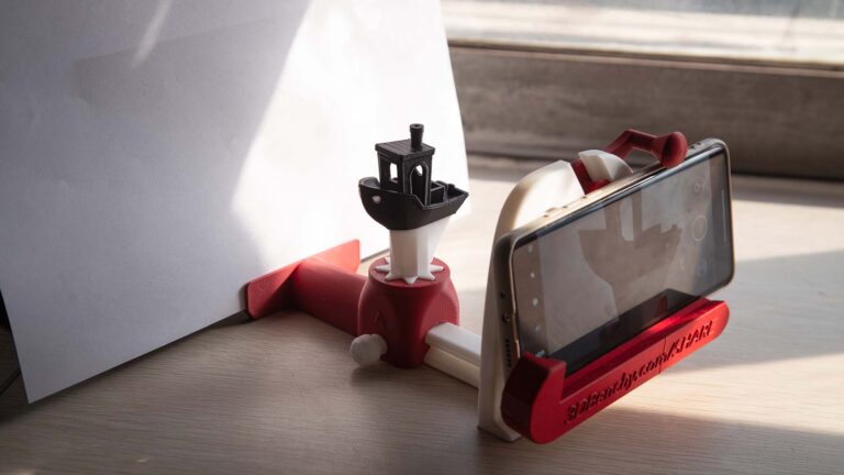 A 3D-printed smartphone camera rig on a wooden surface, featuring a red and white structure holding a smartphone, with a black #3DBenchy model on a rotary platform for 360° video recording, and a sliding fixture for adjusting camera distance and framing.