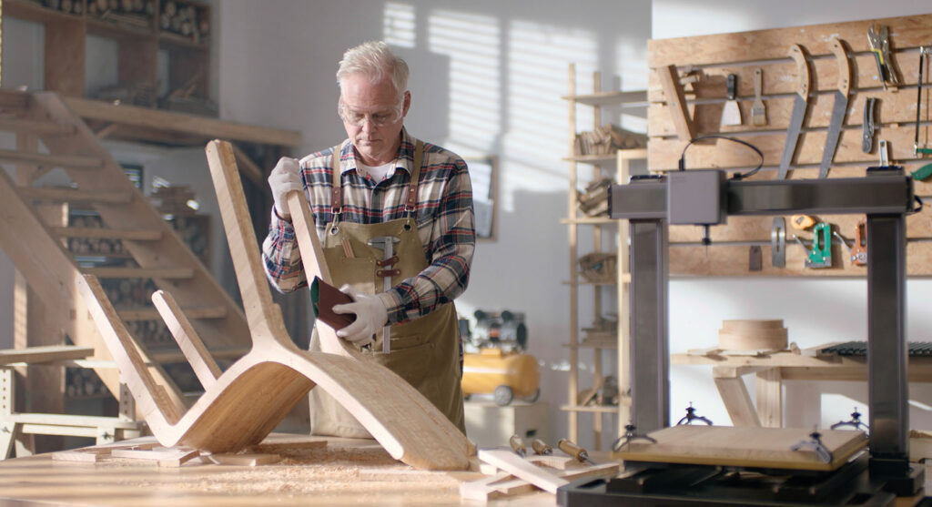 An older man works on a wooden chair frame in a sunlit woodworking shop. He uses tools and wears protective gloves and glasses. The workshop is filled with wooden shelves, a pegboard with various tools, and a large 3D printer or CNC machine in the background, surrounded by sawdust and woodworking equipment.