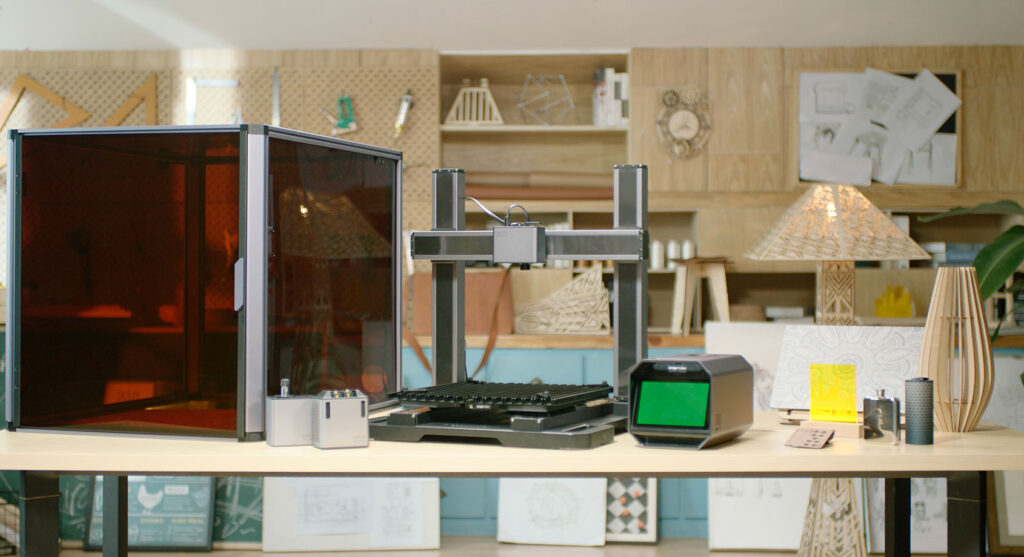 A modern workshop featuring a large 3D printer with a tinted enclosure , positioned on a light wooden table. Surrounding the printer are various accessories, including a small monitor and metallic components.