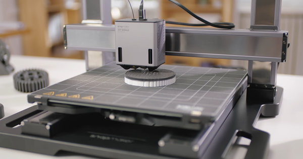 A close-up of a Snapmaker 3D printer actively printing a circular black and white component on its build plate.
